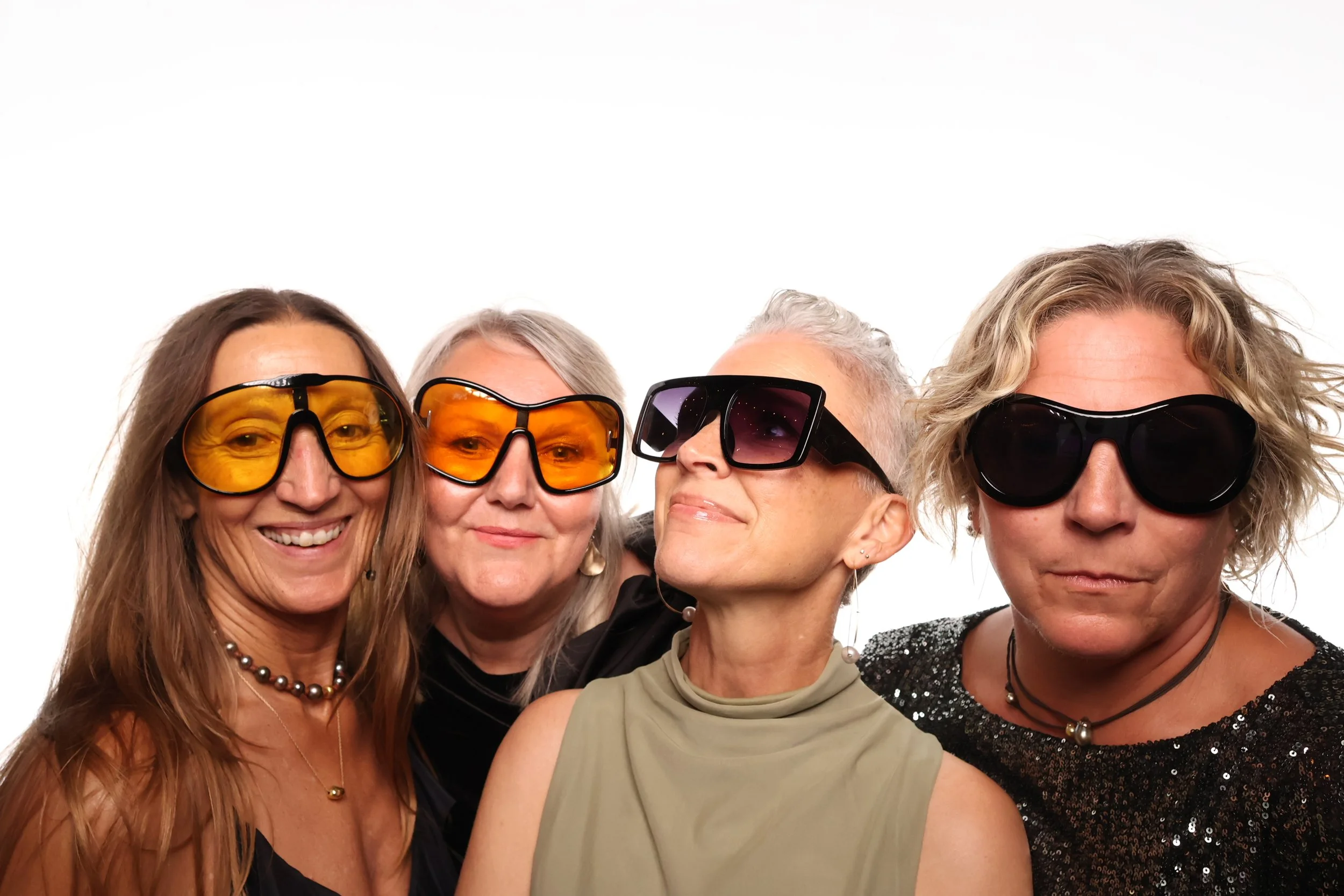 Four women wearing large sunglasses, standing close together against a plain white background.