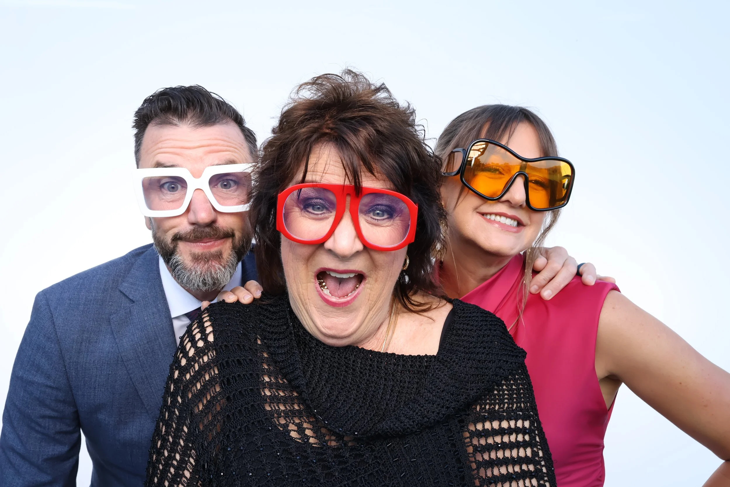 Three joyful people wearing colorful oversized glasses, posing closely together against a plain white background.