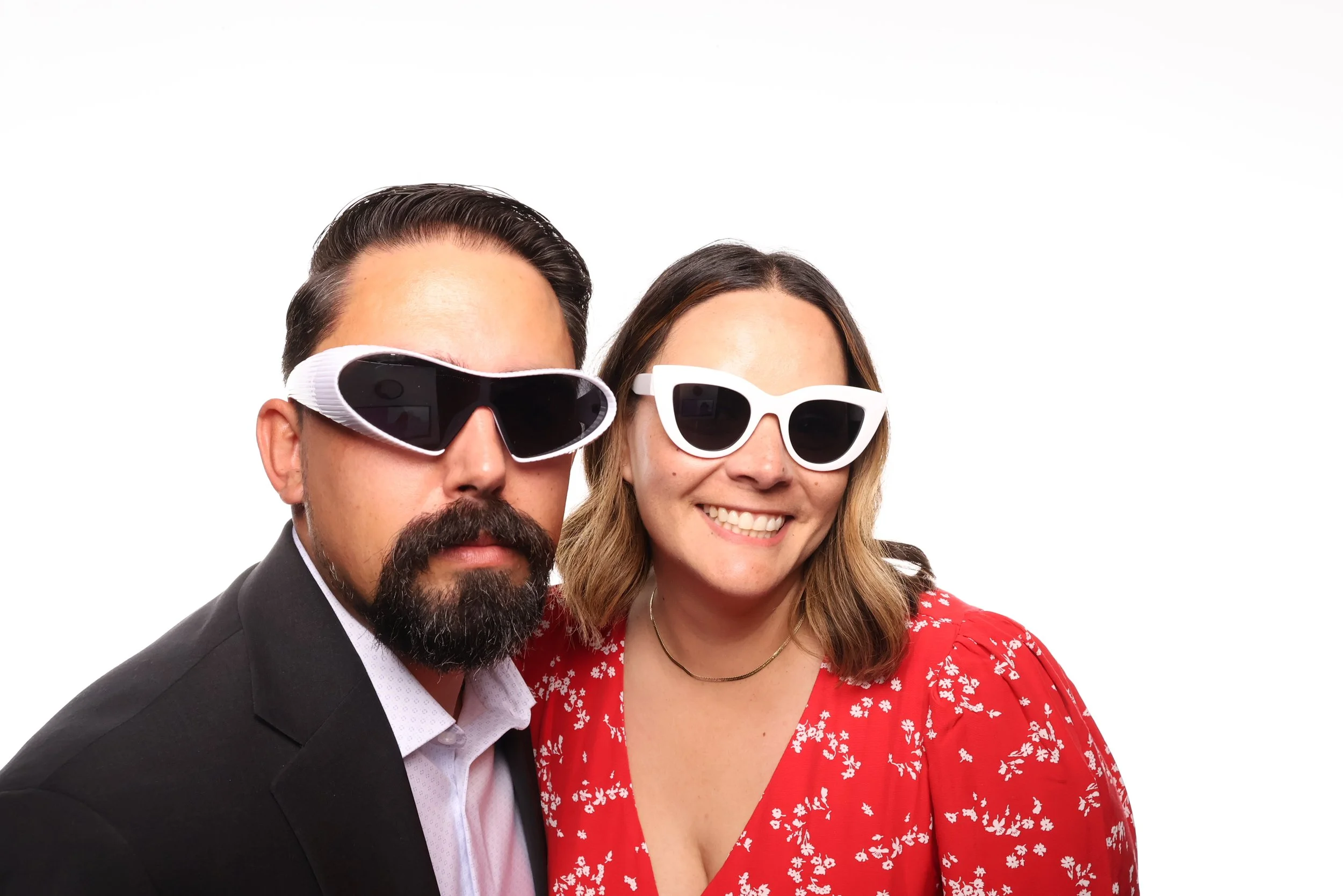 A man and woman wearing sunglasses, smiling, with the woman in a red floral dress and the man in a black suit, against a plain white background.