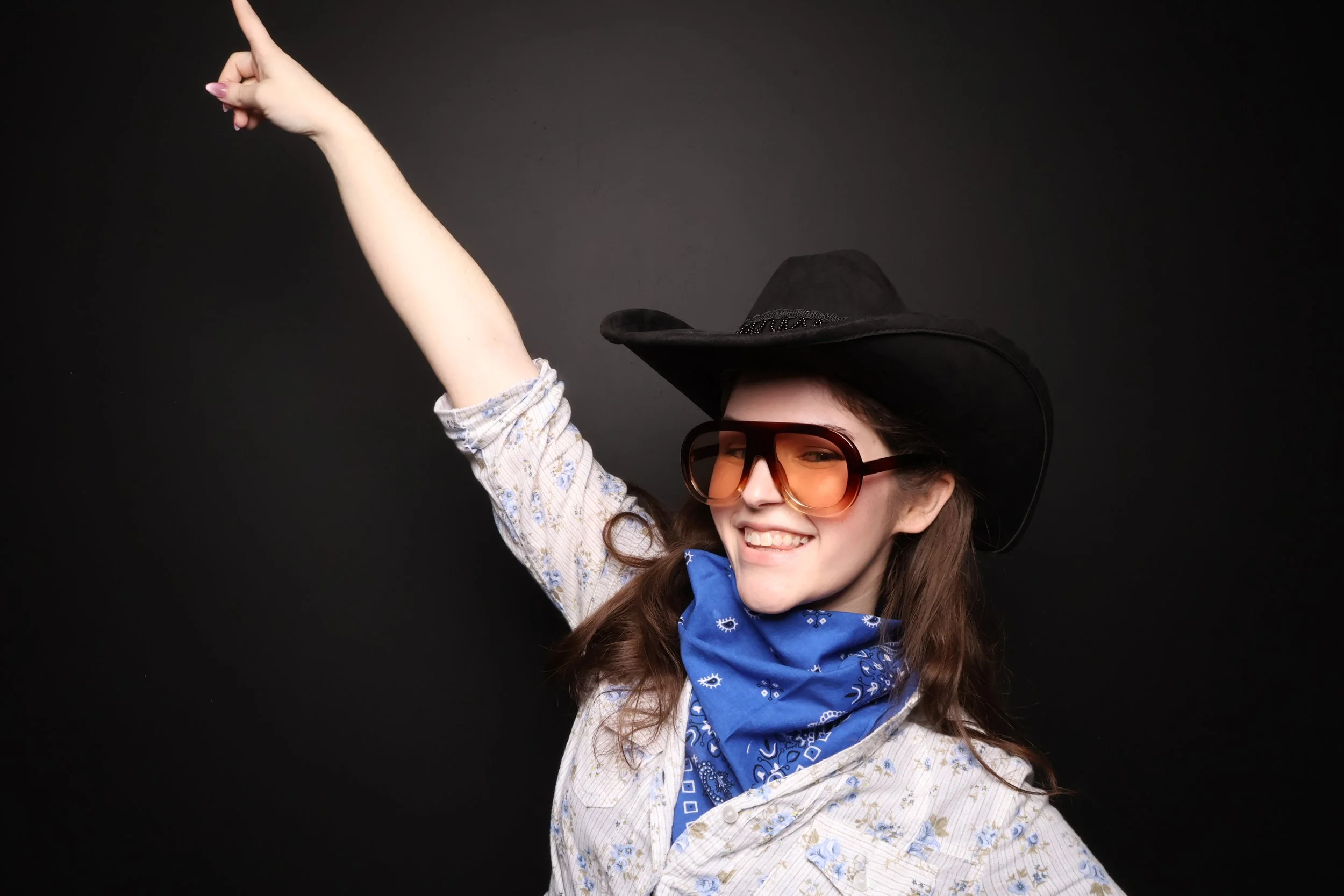 A young woman smiling, wearing a cowboy hat, large sunglasses, a blue bandana around her neck, a white patterned shirt, and a light-colored shirt with blue and purple accents, pointing upward with her right arm against a black background.