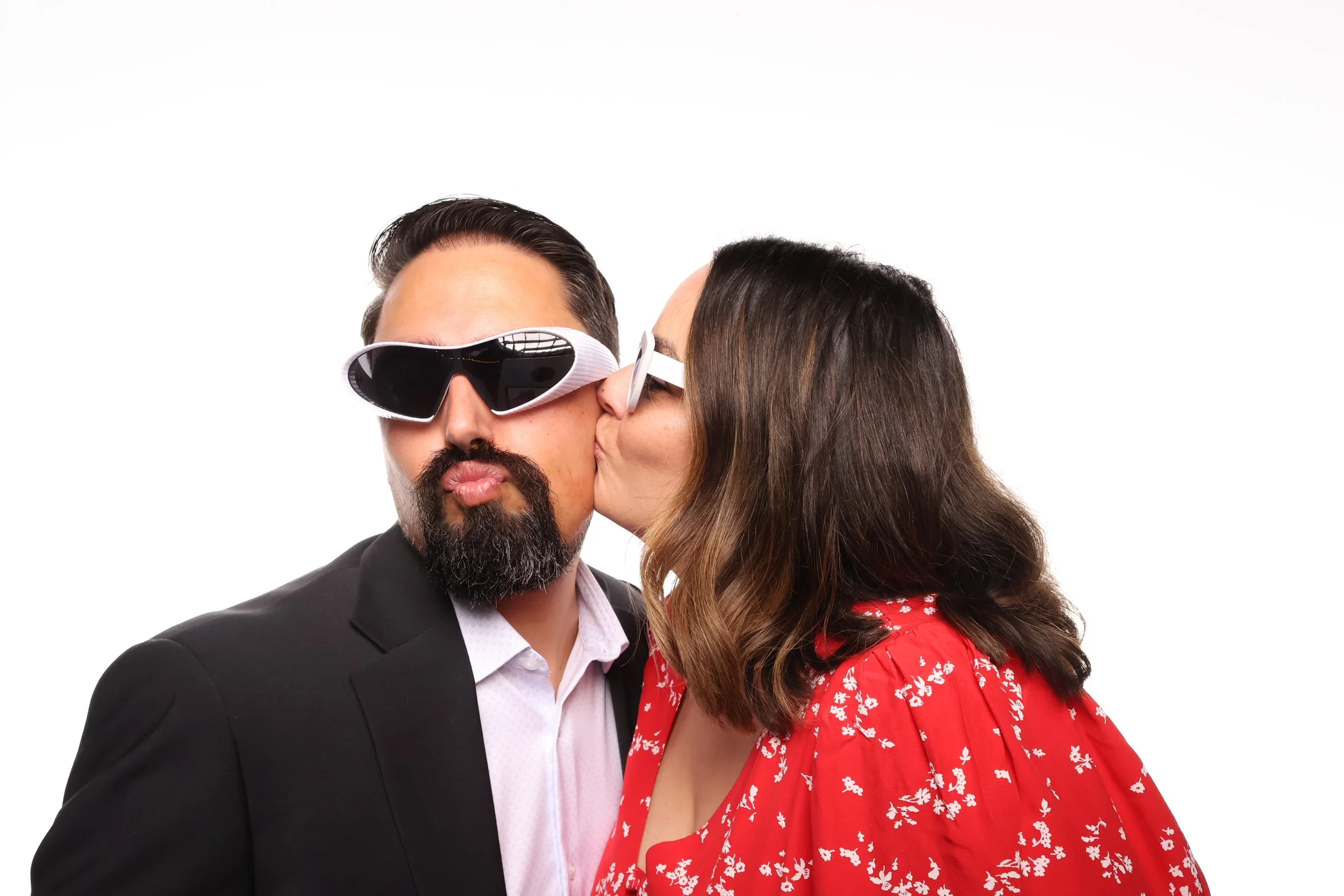 A man with dark hair, a beard, wearing sunglasses and a black suit jacket, is being kissed on the cheek by a woman with long brown hair, wearing a red dress with white floral pattern.