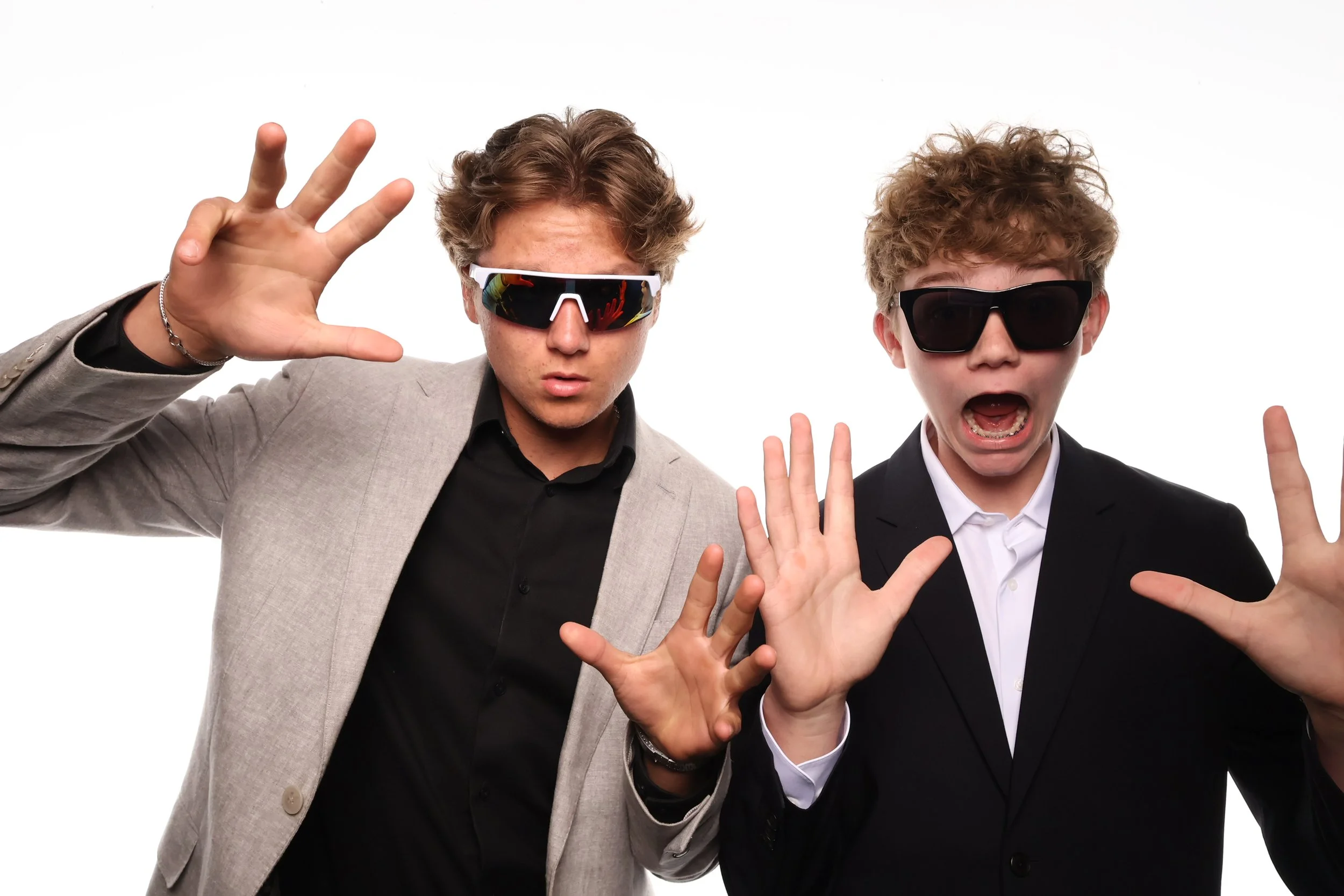 Klixbot fun booth pictures of two young men wearing sunglasses and suits making playful gestures with their hands, standing against a white background.