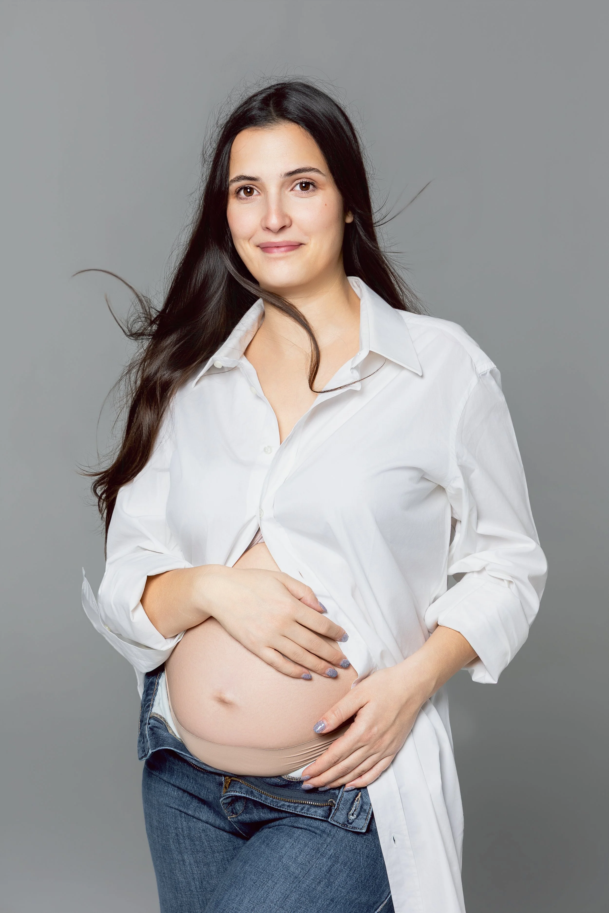 Ritratto fotografico di gravidanza in studio a Milano, con futura mamma in camicia bianca guidata in una posa naturale e delicata.