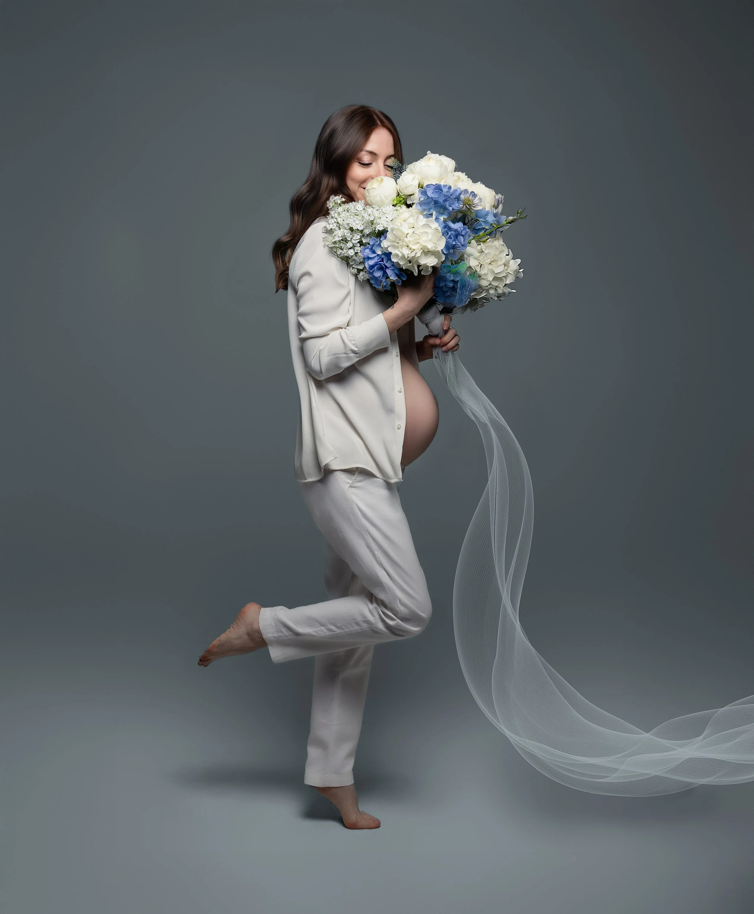 Ritratto fotografico di gravidanza in studio a Milano, con futura mamma vestita di bianco e bouquet di fiori bianchi e azzurri.