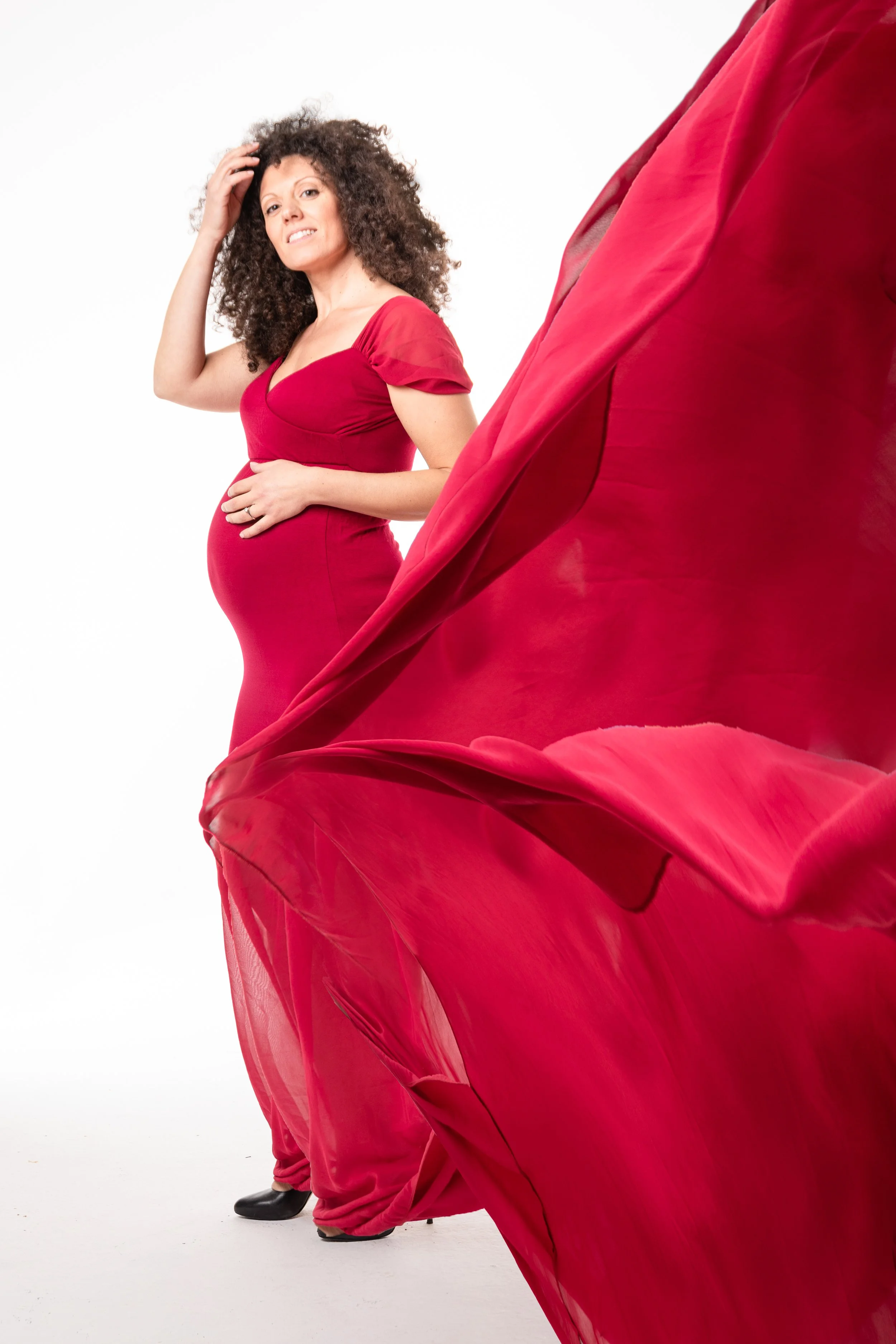 Ritratto fotografico di gravidanza in studio a Milano, con futura mamma in abito rosso e velo coordinato in movimento.