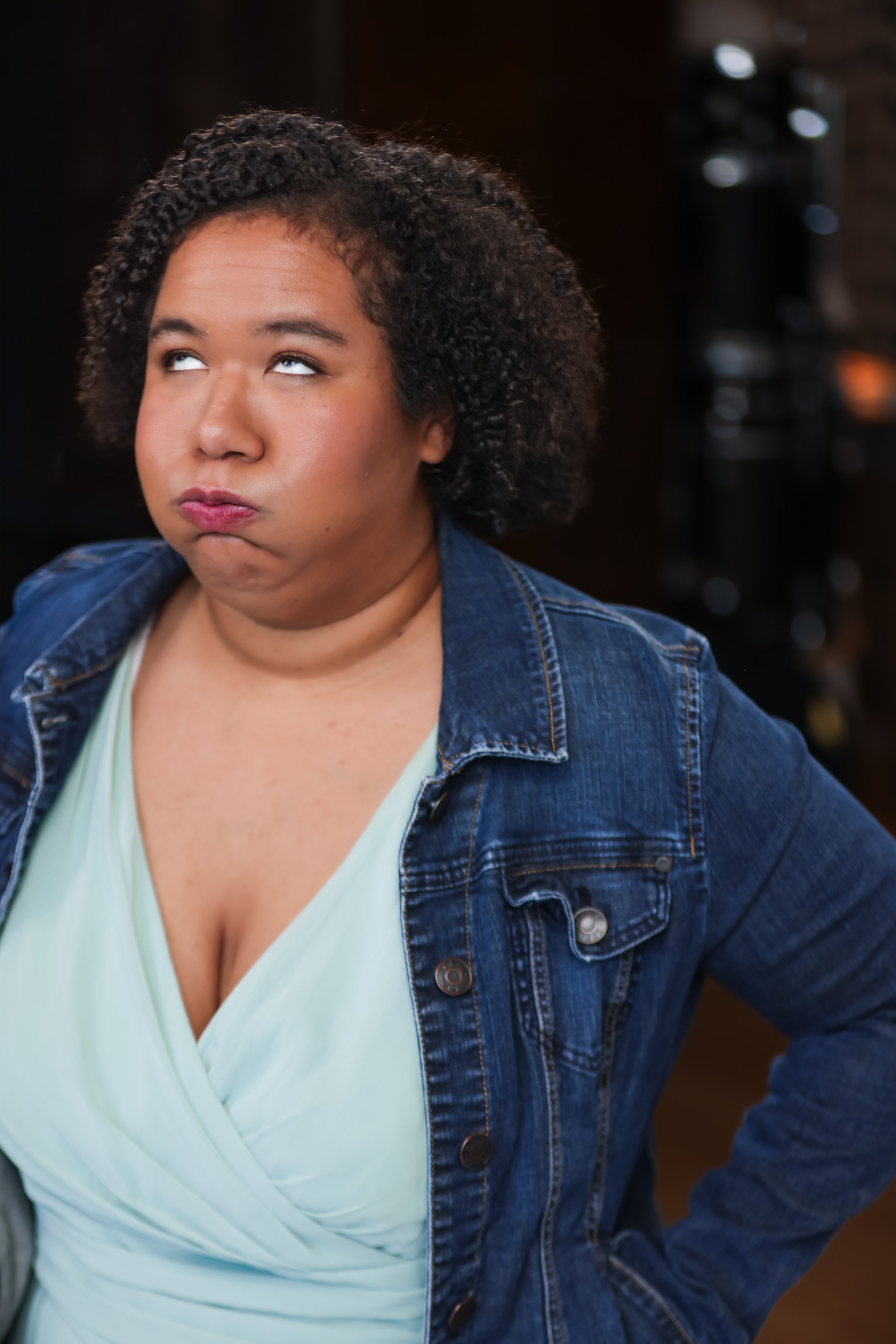 A woman with curly hair wearing a denim jacket and a light green top, looking upward with a puzzled expression.