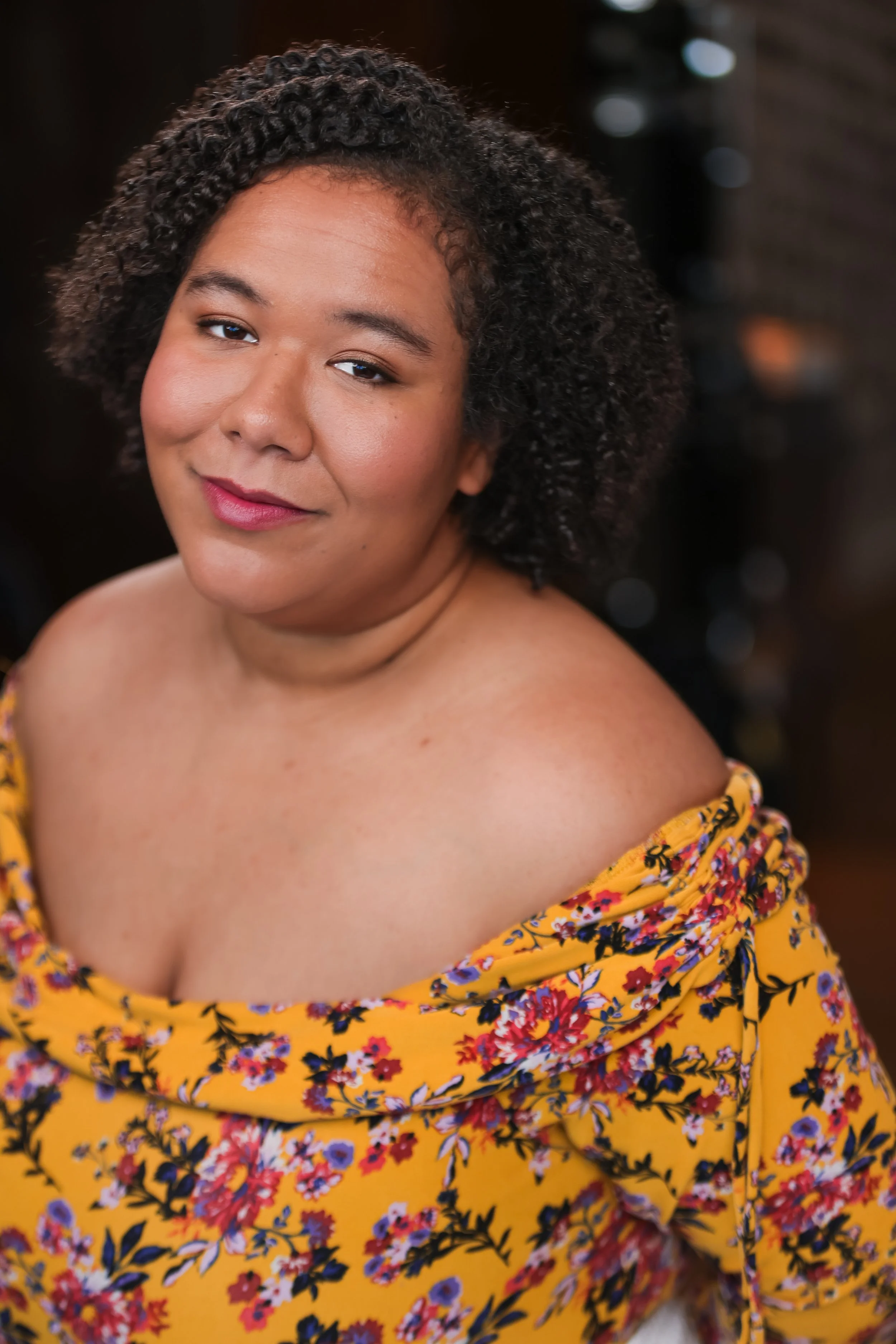 A woman with curly black hair and medium skin, smiling slightly, wearing a yellow floral off-the-shoulder top.