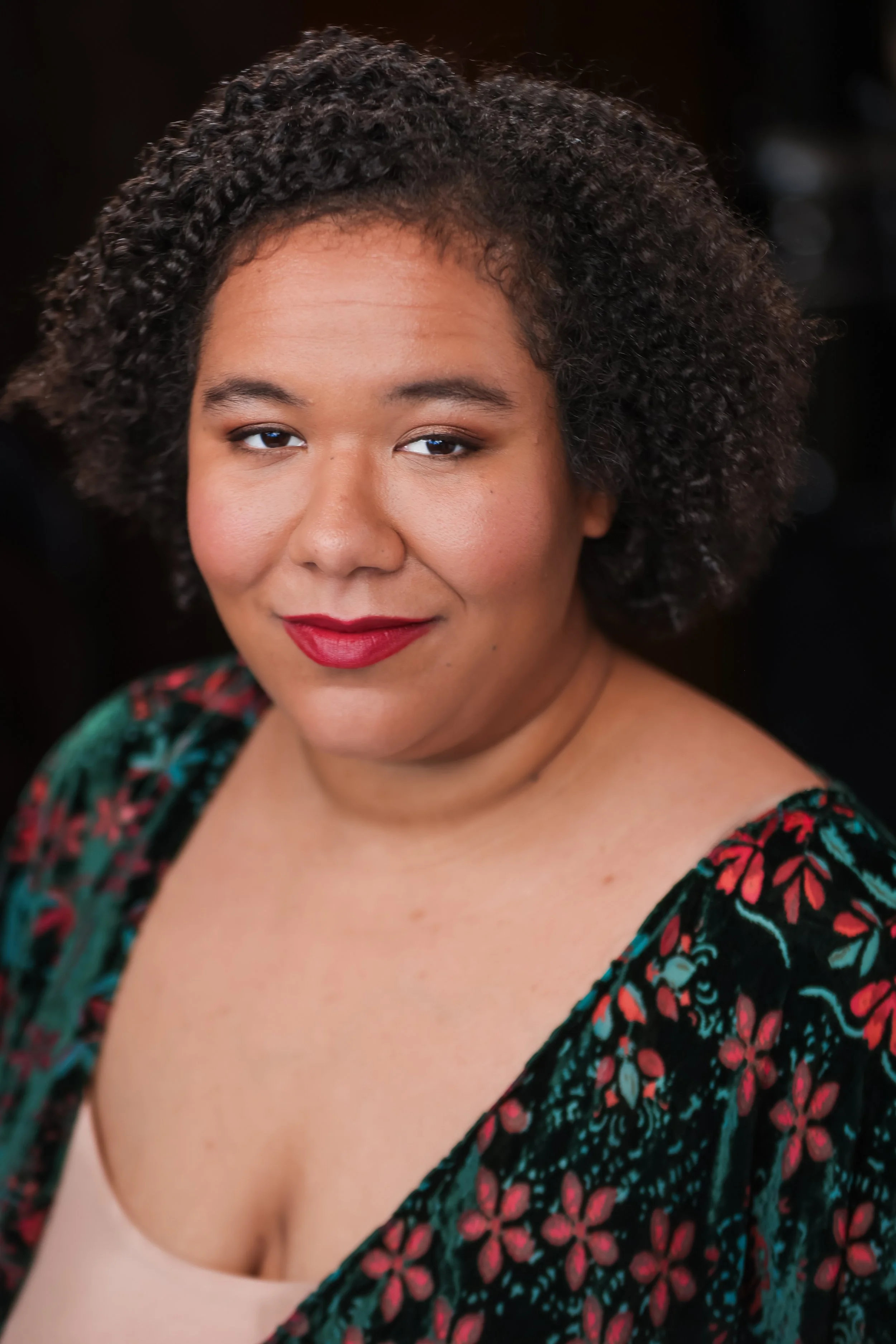 Close-up of a woman with curly hair, wearing red lipstick, in a colorful patterned top with a beige shirt underneath, against a dark background.