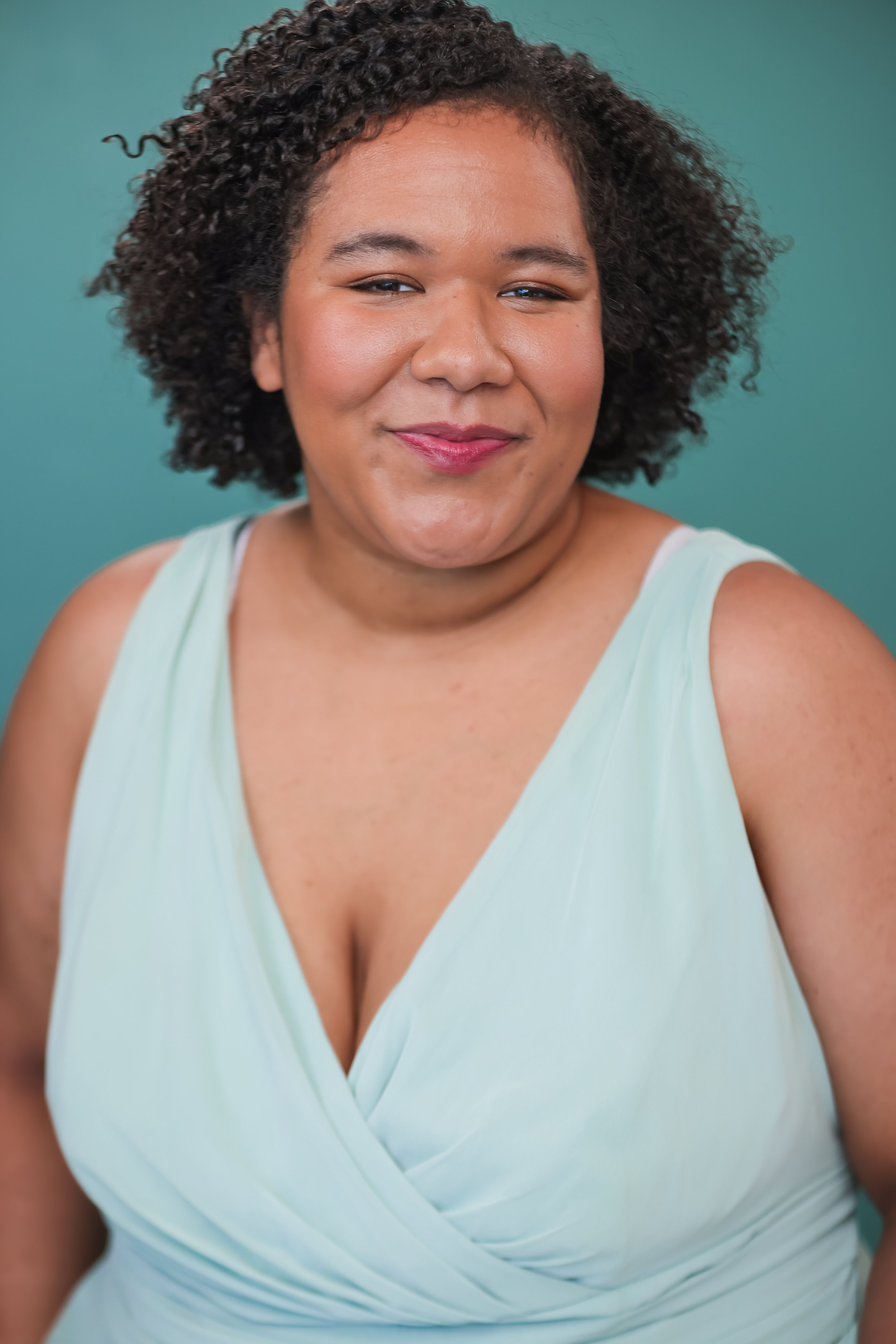 Close-up of a smiling woman with curly hair wearing a light green sleeveless dress against a teal background.