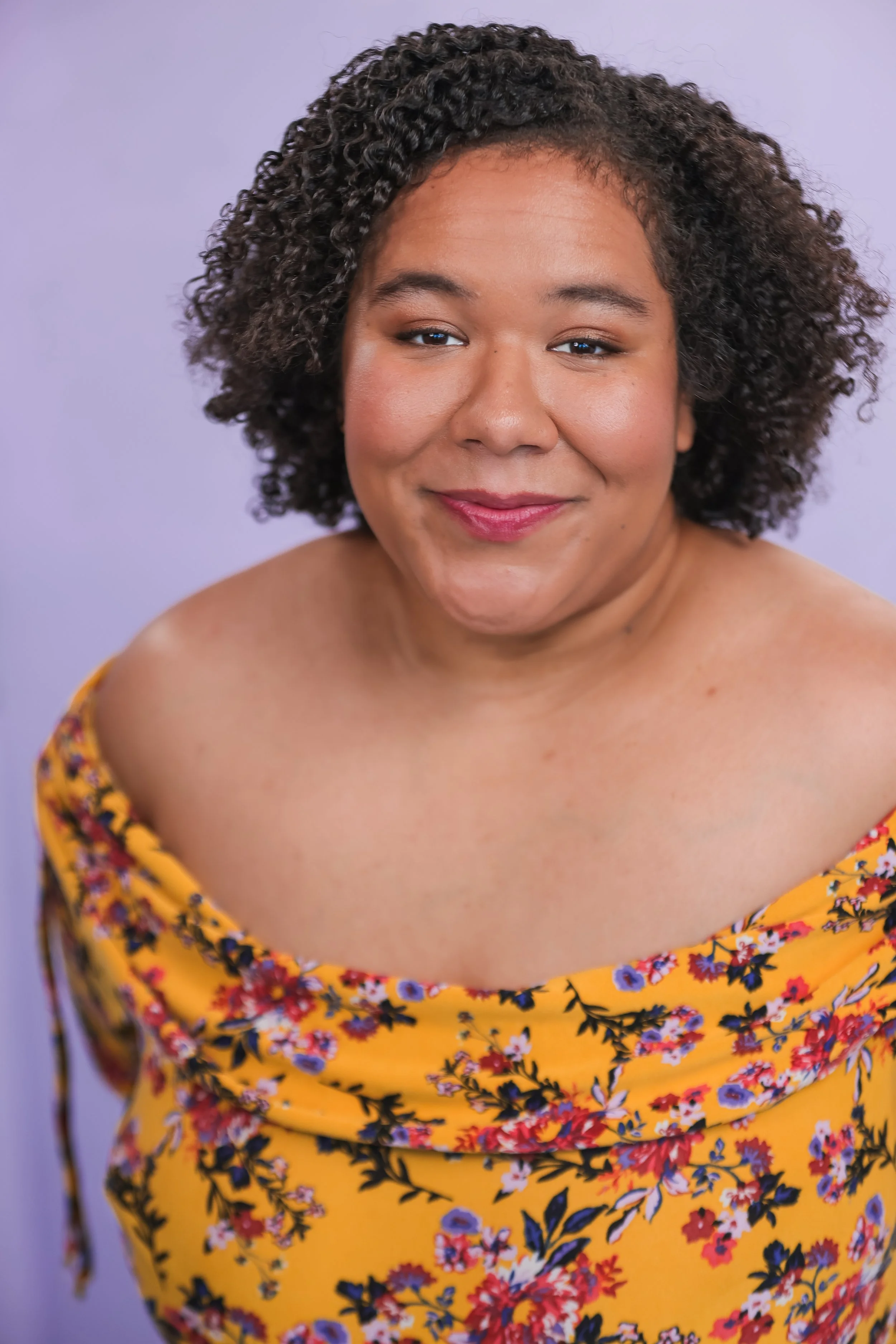 A woman with curly dark hair and a warm complexion is smiling softly. She is wearing a yellow dress with a colorful floral pattern and is set against a plain, light purple background.