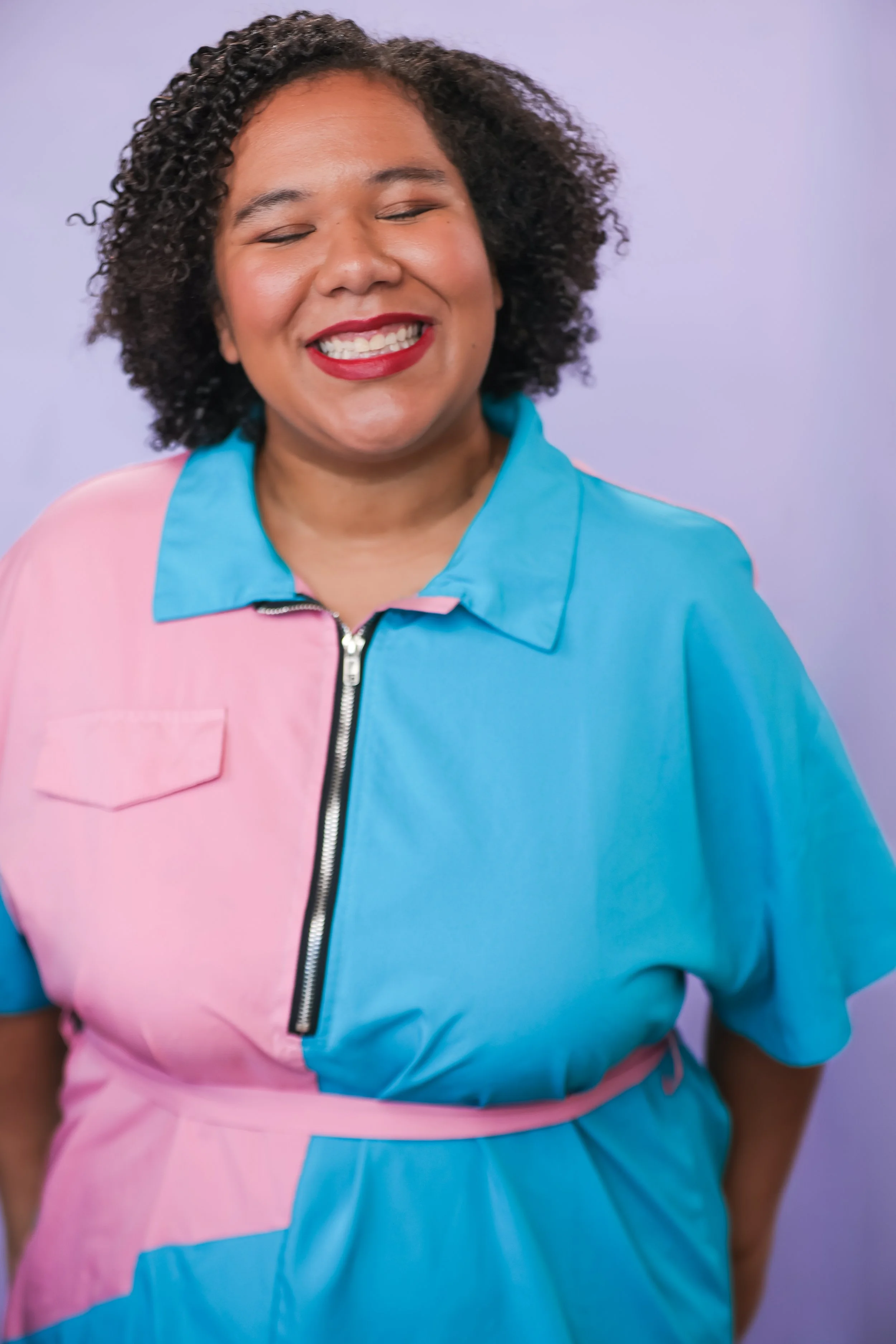 A woman with curly hair smiling with red lipstick, wearing a color-blocked jacket with pink and blue sections, set against a light purple background.