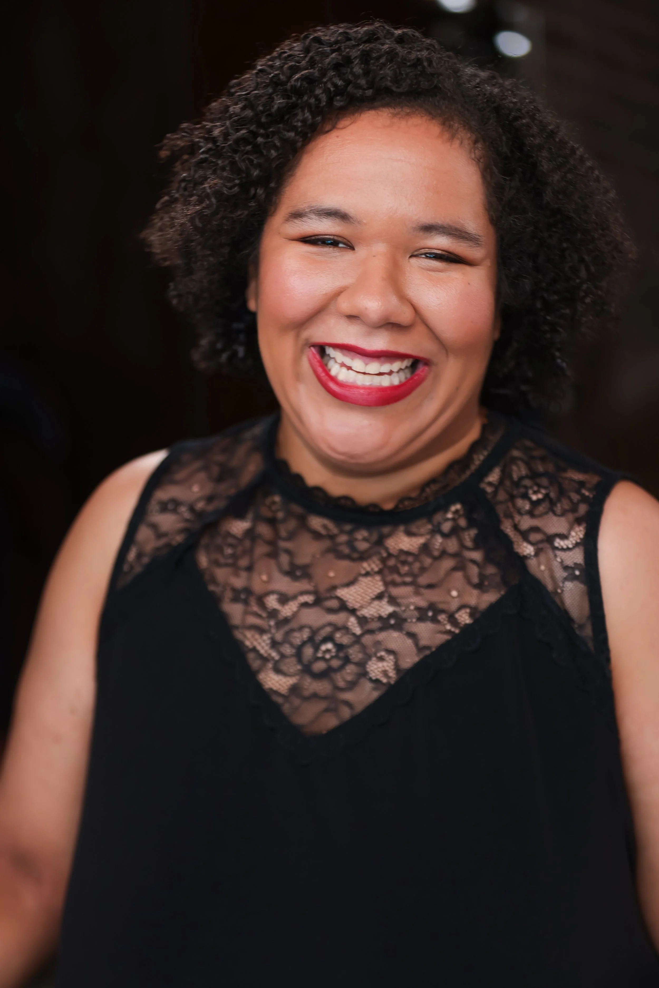 A woman smiling widely, showing her teeth, with curly black hair, wearing a black lace top.