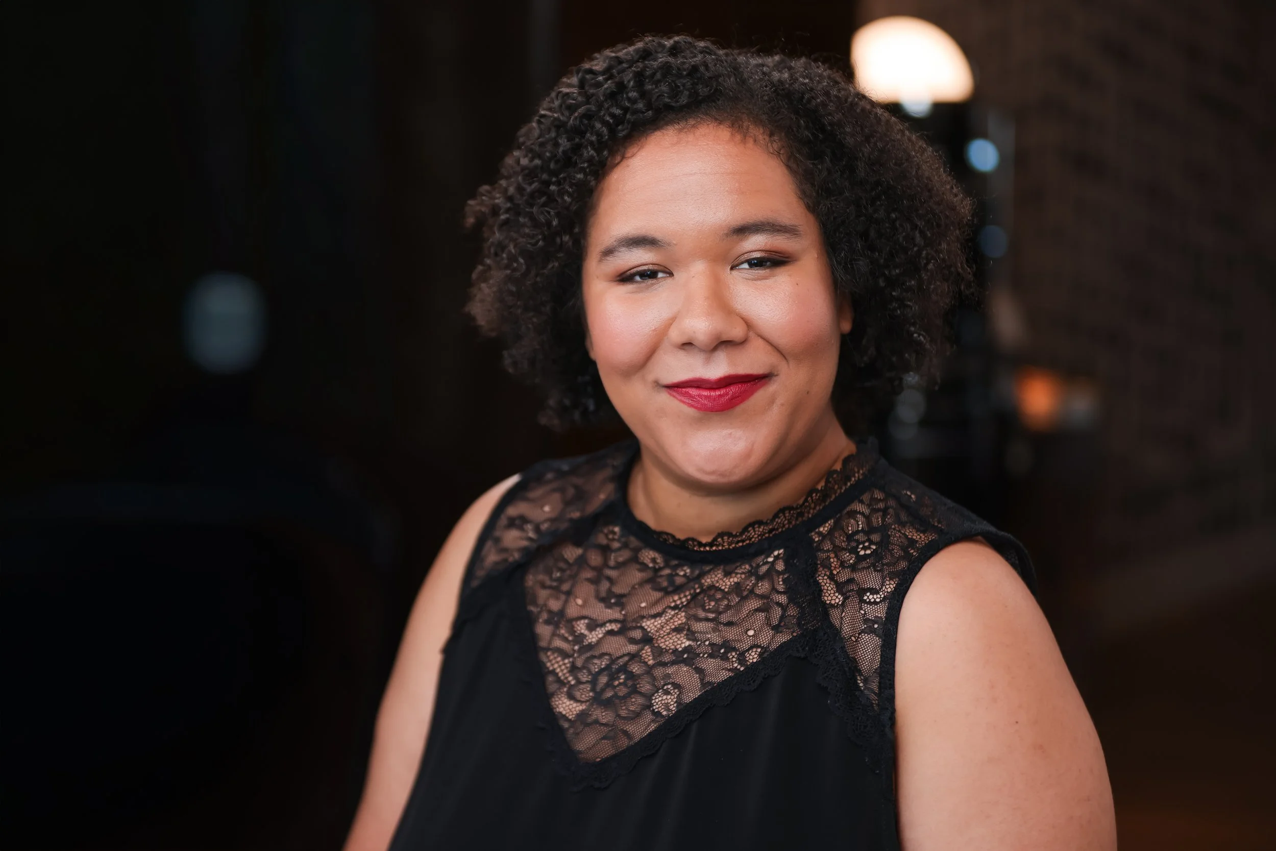 A woman with curly hair and red lipstick wearing a black lace dress, smiling gently, in a dimly lit indoor setting.