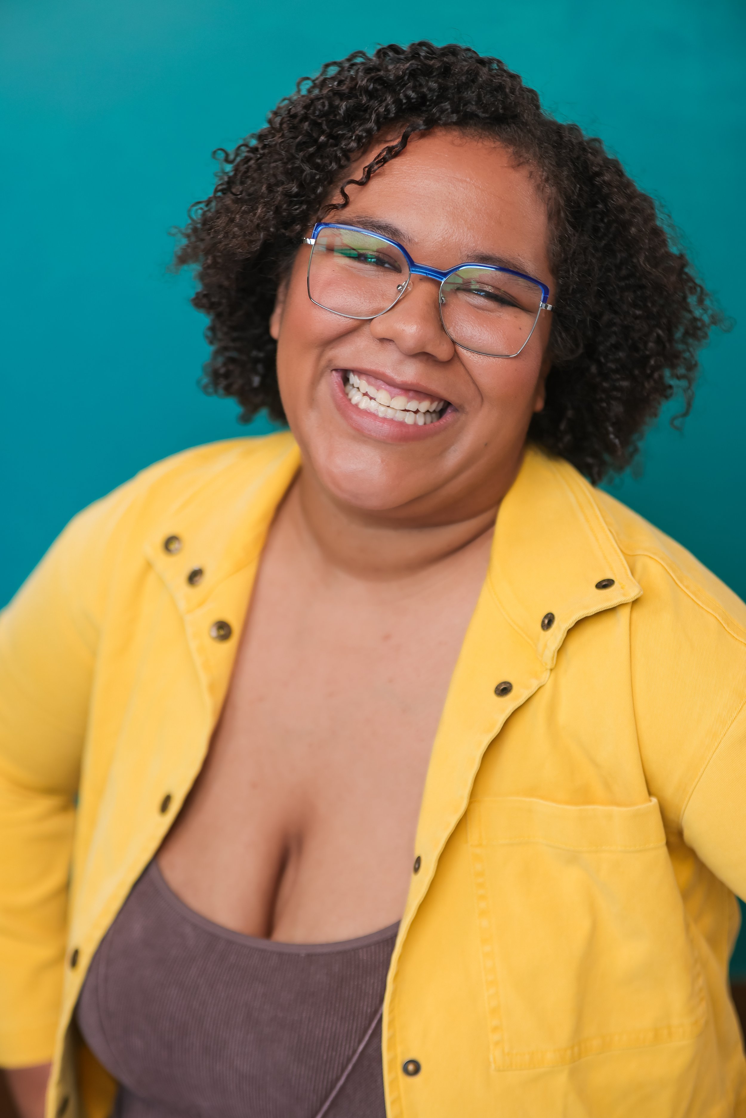 A woman with curly dark hair, wearing glasses with blue frames, a yellow jacket, and a brown top, smiling against a teal background.