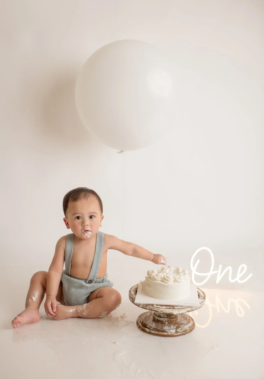 A young child with some icing on his face sitting on the floor next to a white frosted cake on a stand, with a large white balloon behind him and the word "One" written in cursive to the right of the cake.