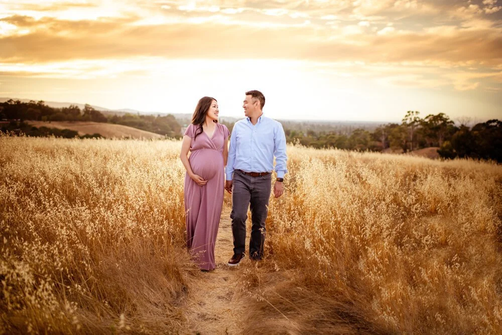 A pregnant woman and a man walking through a field of tall grasses during sunset, holding hands and smiling at each other.