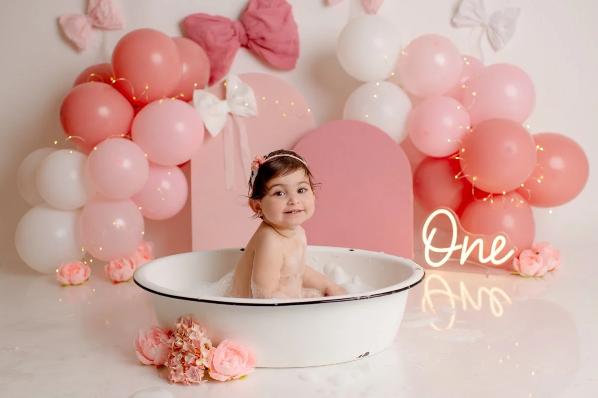 A young girl in a bathtub surrounded by pink and white balloons, pink flowers, and a pink backdrop with the word 'One' glowing in the foreground, celebrating her first birthday.