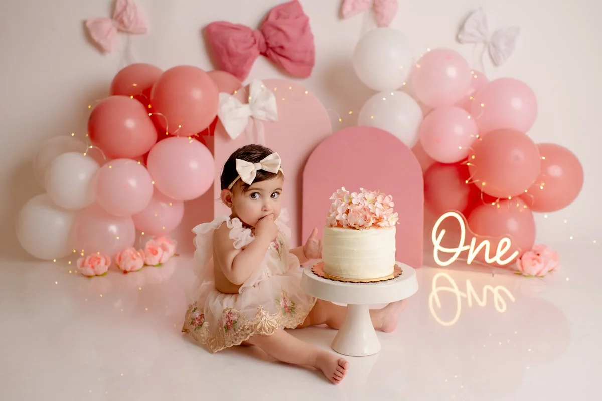 A young girl with a bow in her hair celebrating her first birthday, sitting beside a cake with pink and white decorations, surrounded by pink and white balloons and bows, with a neon sign that says 'One' next to her.