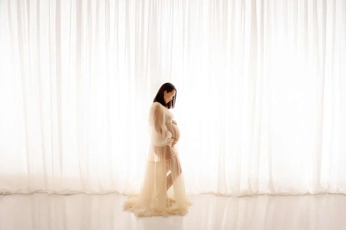 A pregnant woman in a light, flowing dress standing in front of white curtains, softly smiling and looking down at her belly.