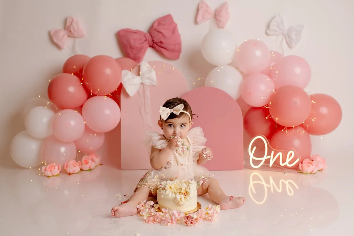 A young girl celebrating her first birthday with a cake, surrounded by pink and white balloons, bows, and flowers in a pastel-colored setup.