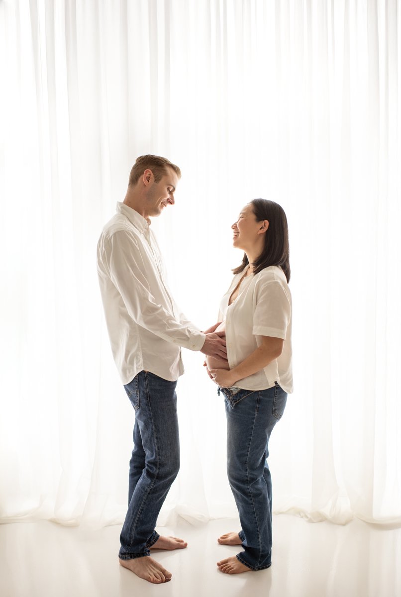A couple standing barefoot in front of sheer white curtains, with the woman holding her pregnant belly and the man gently touching her belly, smiling at each other.
