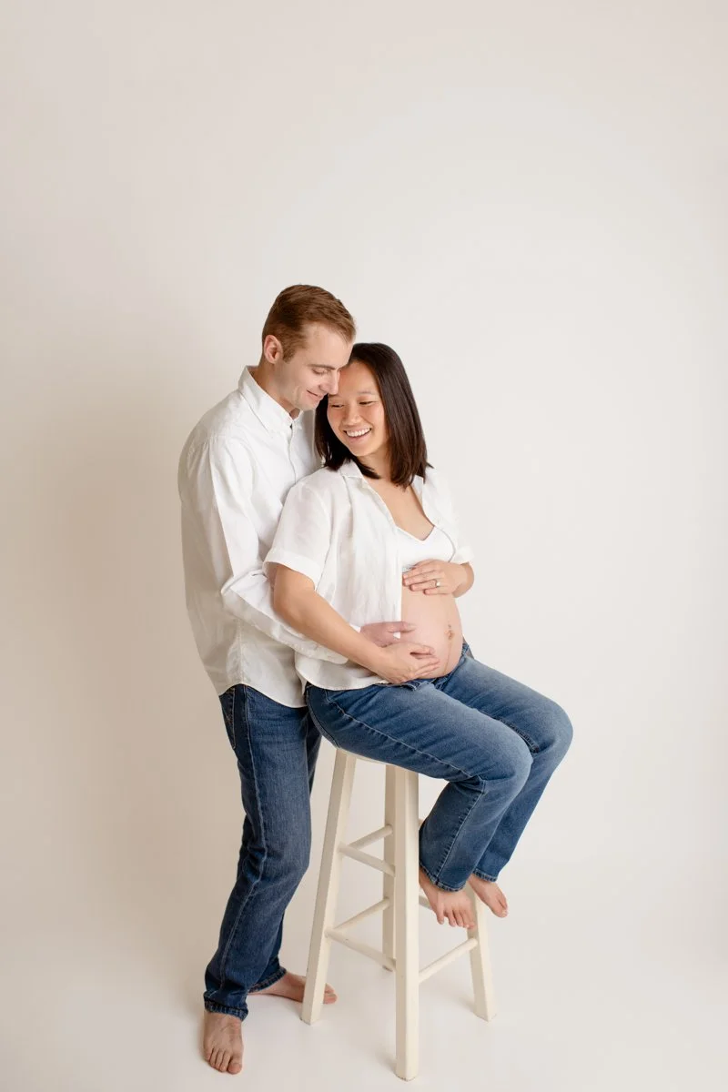 Pregnant woman sitting on a stool with her partner standing behind her, both smiling and touching her belly.