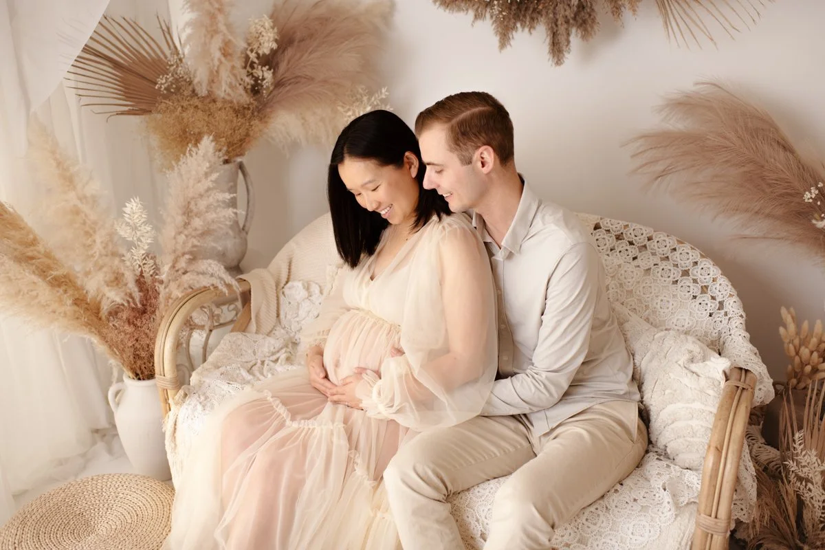 A pregnant woman sitting on a wicker couch with her partner, both smiling and looking at her belly. The scene is decorated with beige pampas grass and dried flowers in a white vase.
