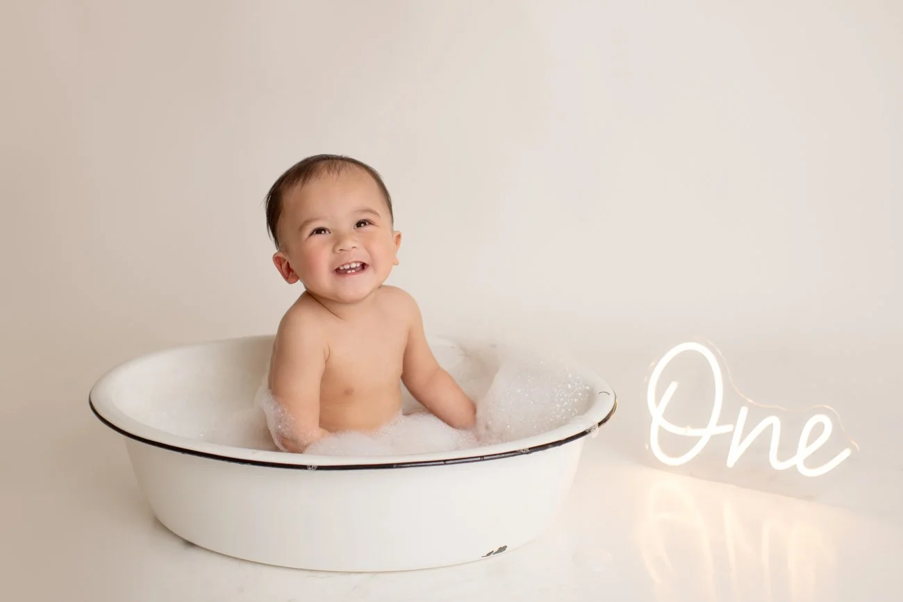 A smiling baby sitting in a white oval bathtub filled with bubbles, with a neon sign that says "One" on the right side.