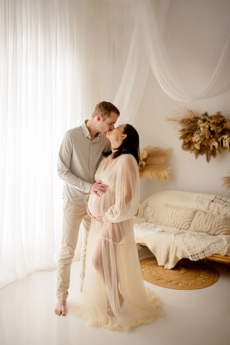 A pregnant woman and a man are sharing a kiss in a softly lit, cream-colored room with bohemian decor, including dried floral wall arrangements, sheer curtains, and cozy woven furniture.