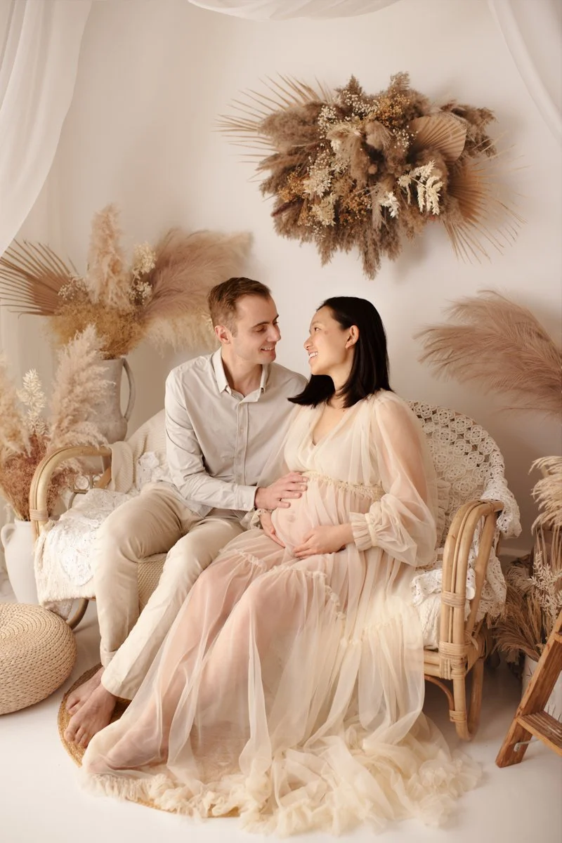 A couple sitting on a wicker chair, with the woman pregnant, surrounded by pampas grass and dried flowers in a neutral-toned setting.