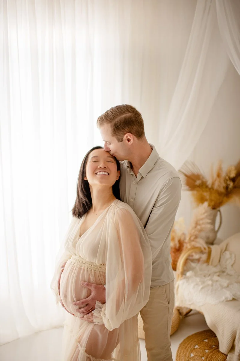 A happy pregnant woman with dark hair and a big smile, standing in a sunlit room, with a man kissing her forehead. The man has light hair and is embracing her from behind. The woman is wearing a sheer, flowing dress, and the room is decorated with wh