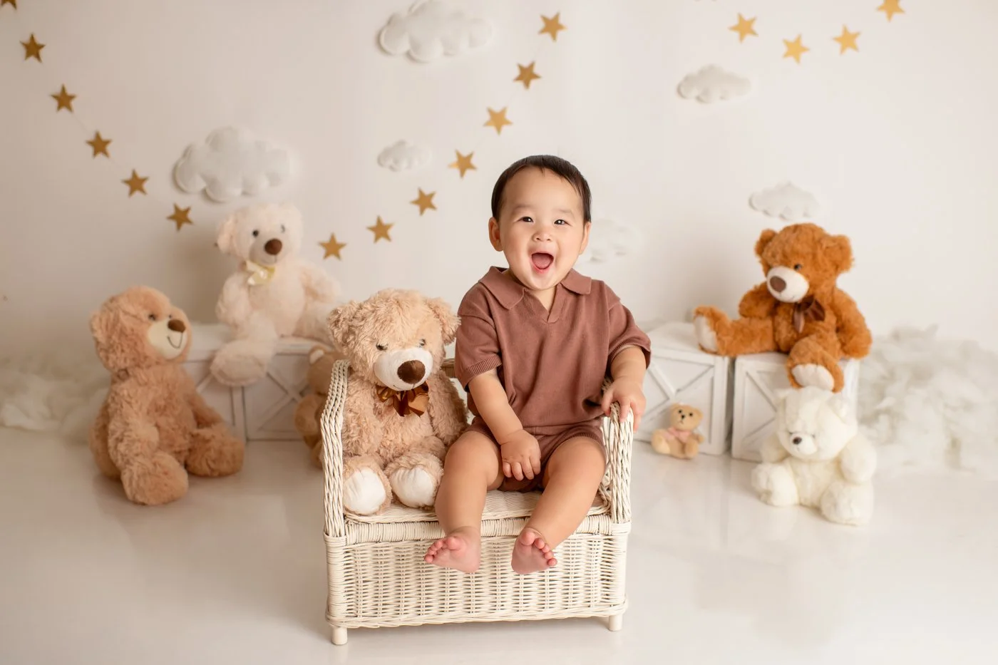 Smiling young child sitting on a small wicker chair surrounded by various teddy bears in a decorated room with cloud and star wall art.