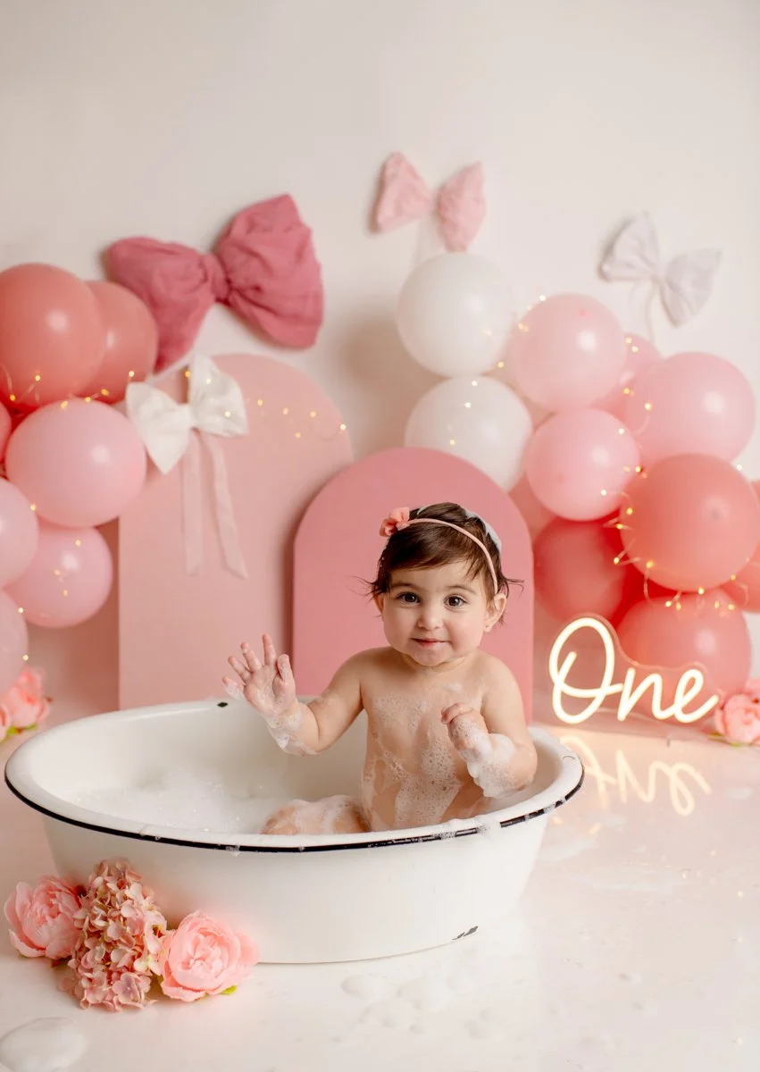 A young girl taking a bath in a white tub surrounded by pink, white, and peach flowers, with pink and white balloons and bows in the background celebrating her first birthday.