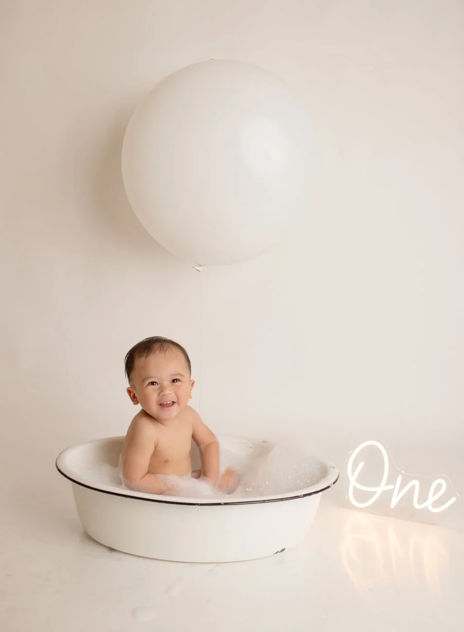 A happy baby sitting in a white bath tub with bubbles, a large white balloon hanging above, and a neon sign that reads 'One' on the floor beside the tub.