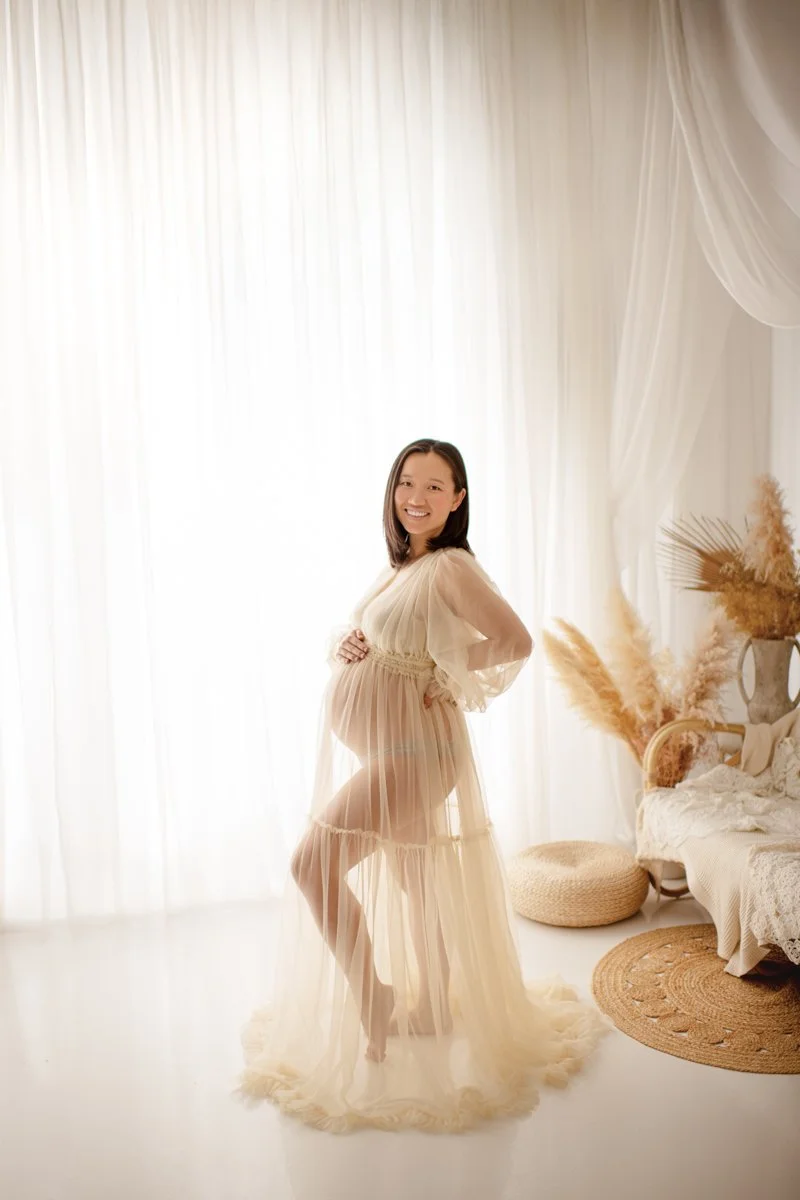 A pregnant woman smiling in a beige sheer dress standing indoors with a bright, airy background, next to a bed with rustic beddings and dried pampas grass in vases.