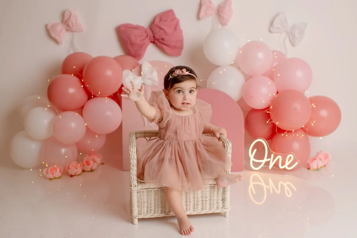 A young girl in a peach-colored dress sitting on a wicker chair, surrounded by pink and white balloons, with decorative bows and flowers in the background, celebrating her first birthday.