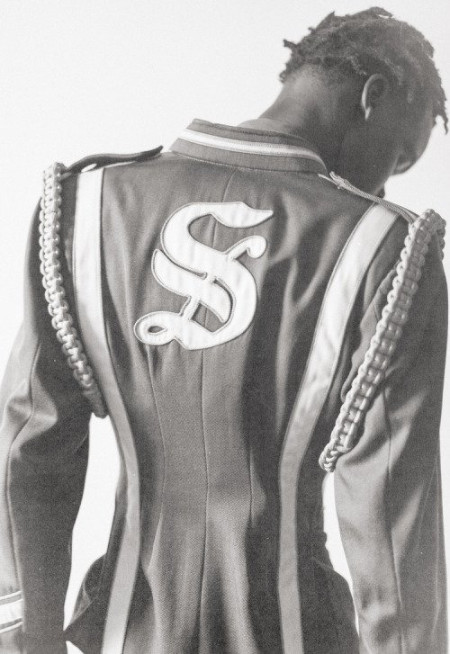 Black and white photo of a person wearing a decorative jacket with a stylized 'S' on the back, showing long braids or dreadlocks. Portrait by Anthony Amadeo