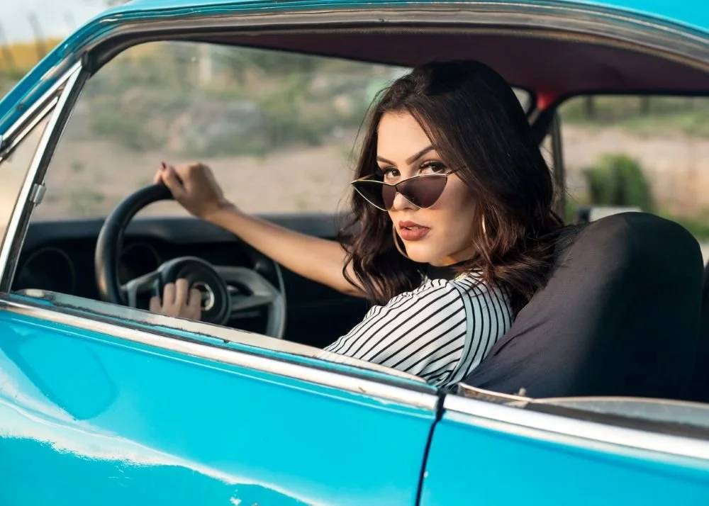woman sitting in car looking back at camera with sunglasses lowered