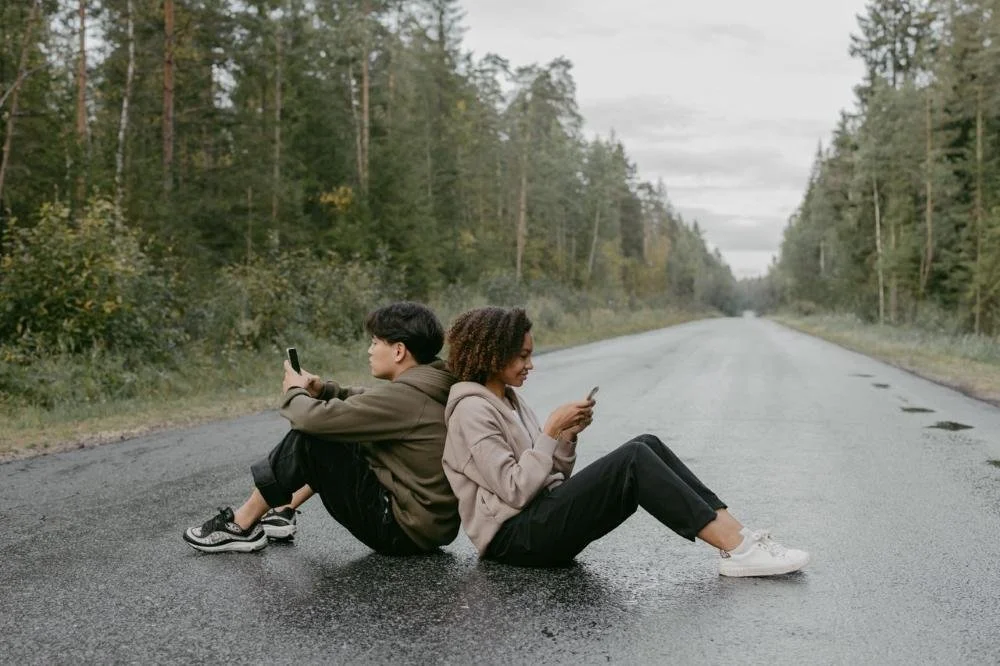 a couple sitting with backs against each other on a road looking at cellphones