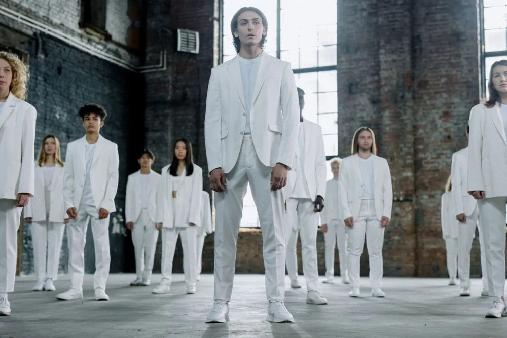 A diverse group of models in elegant white suits standing in an industrial indoor setting.
