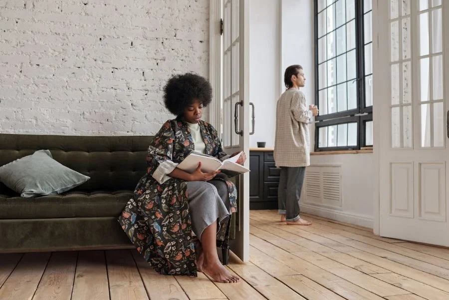 a woman sitting in one room reading a book and a male in another looking out a window
