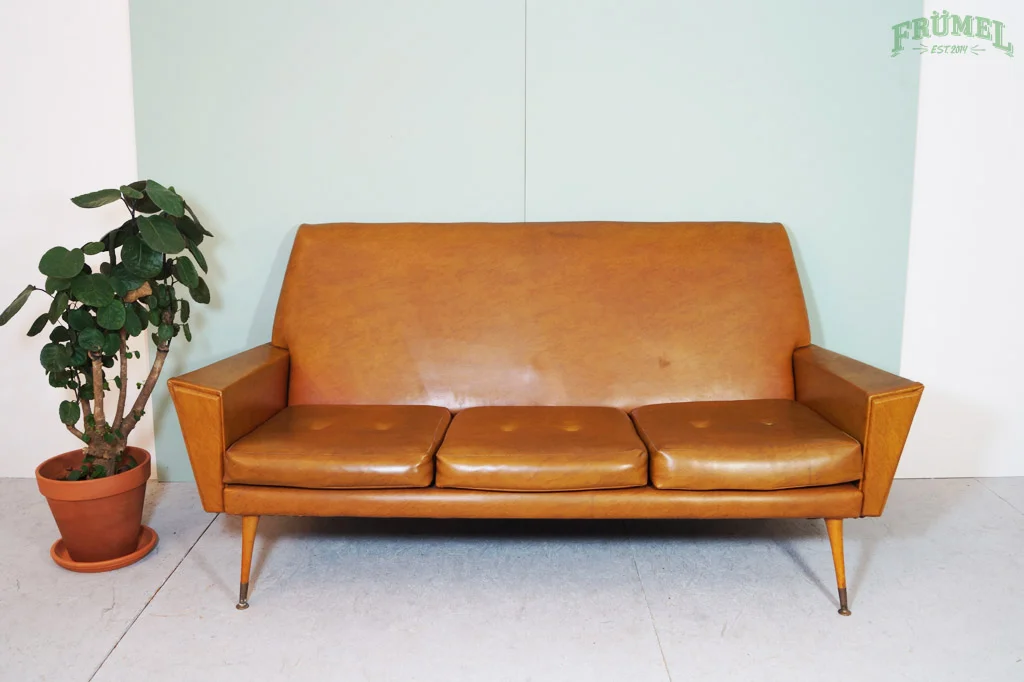 Een vintage bruine leren bank met drie zitplaatsen naast een grote groene plant in een terracotta pot, tegen een lichte muur met een groene tekst logo op de rechterboven hoek.