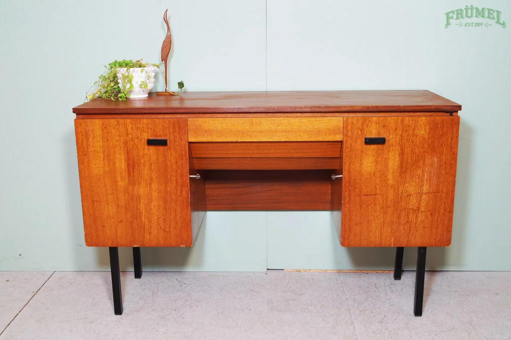 Een houten bureau met zwarte poten en een plant op de linkerzijkant, tegen een lichtblauwe muur.