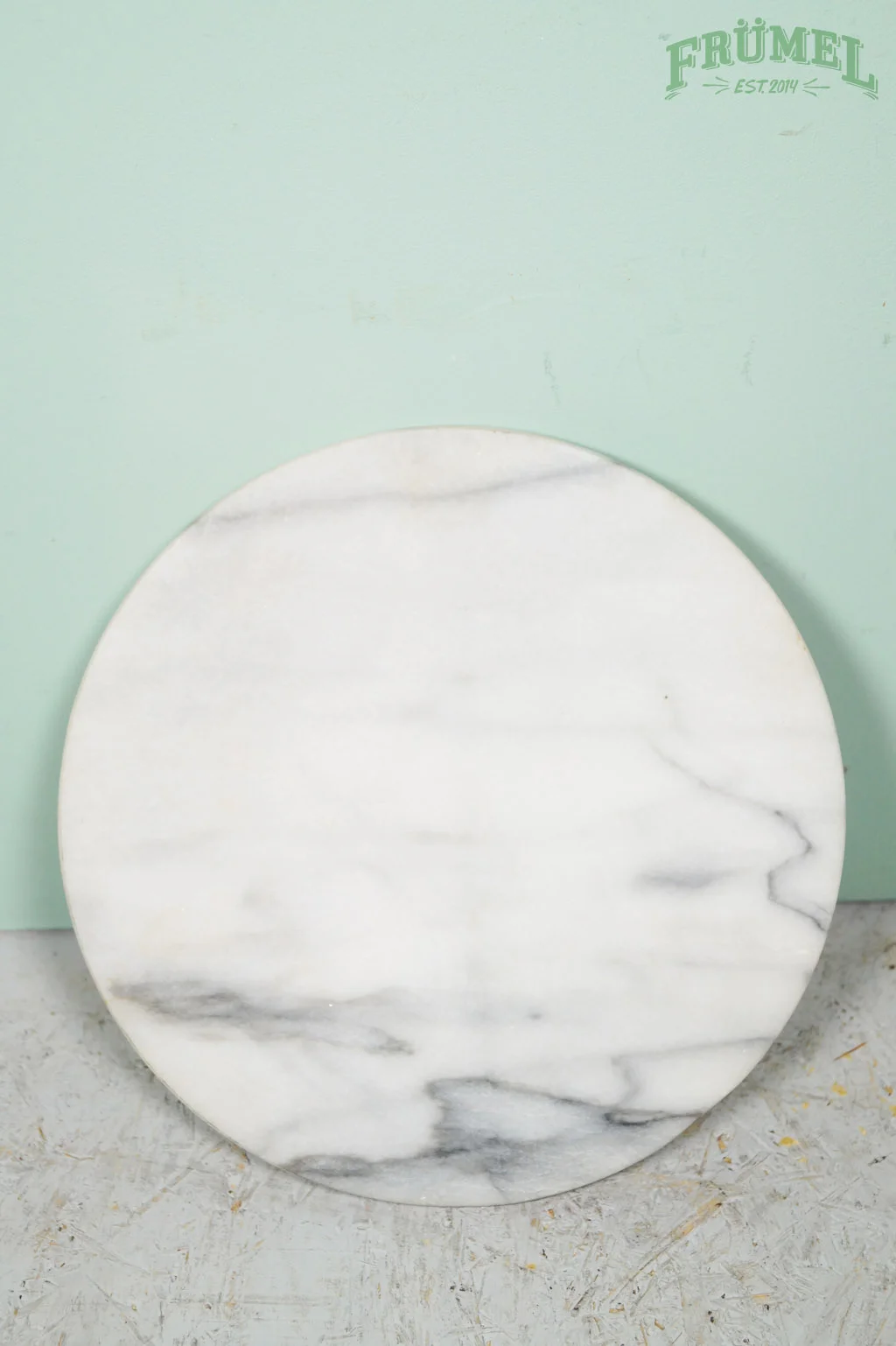 Een ronde, witte marmeren snijplank met grijze aderen, geplaatst op een betonlook vloer, met een groene muur op de achtergrond en een logo 'FRÜMEL EST. 2014' in de rechterbovenhoek.