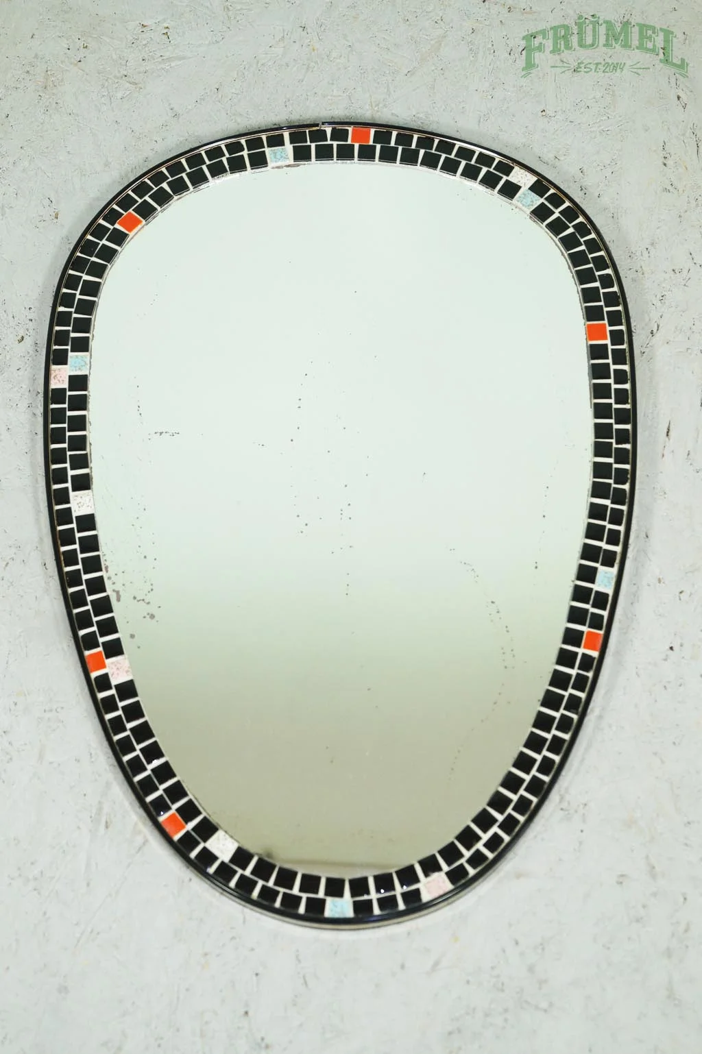 Muurspiegel met zwarte en oranje kader, op een lichte muur met de tekst 'Frümel' en 'EST 2014' erboven.