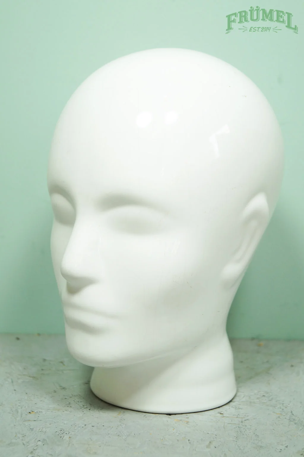 Witte barbie-hoofd mannequin met gesloten ogen en zonder haar op een grijze ondergrond, groene achtergrond en een logo "FRÜHEL" in de rechterbovenhoek.