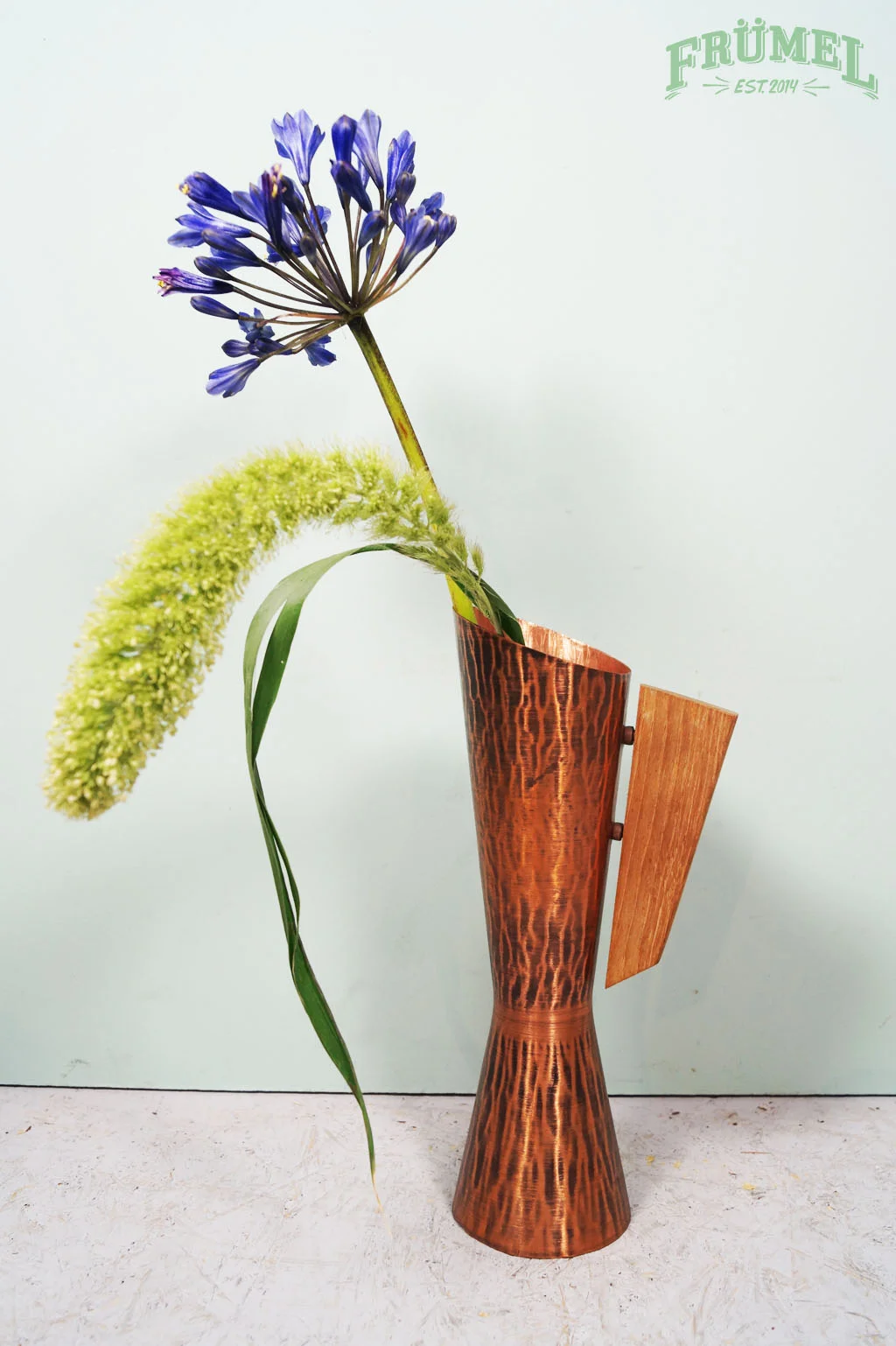 Een vaas met een blauwe allium bloem, een witte bloempluim en een groene blad. De vaas is gemaakt van metaal en hout.