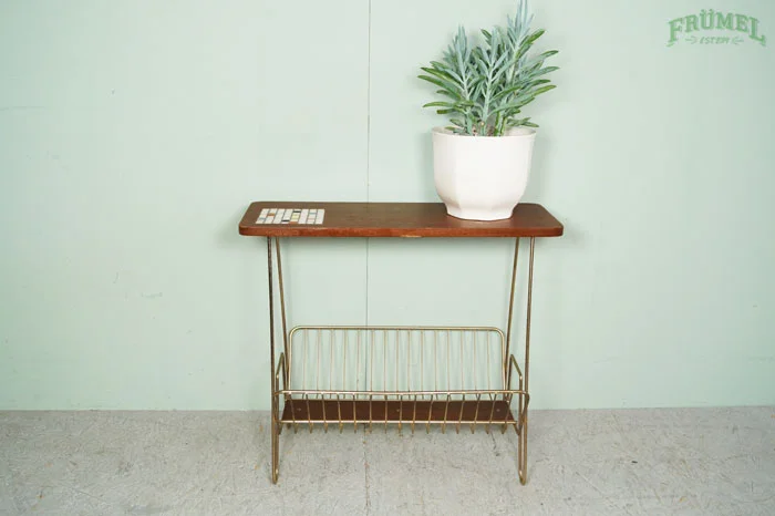 Een groene plant in een witte pot op een houten tafel met een metalen rek eronder, tegen een lichtgroene muur met een groen logo.