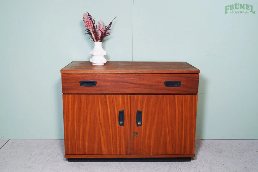 Houten kast met twee deuren en een lade, met een witte vaas met bloemen erop, tegen een lichte groene muur.