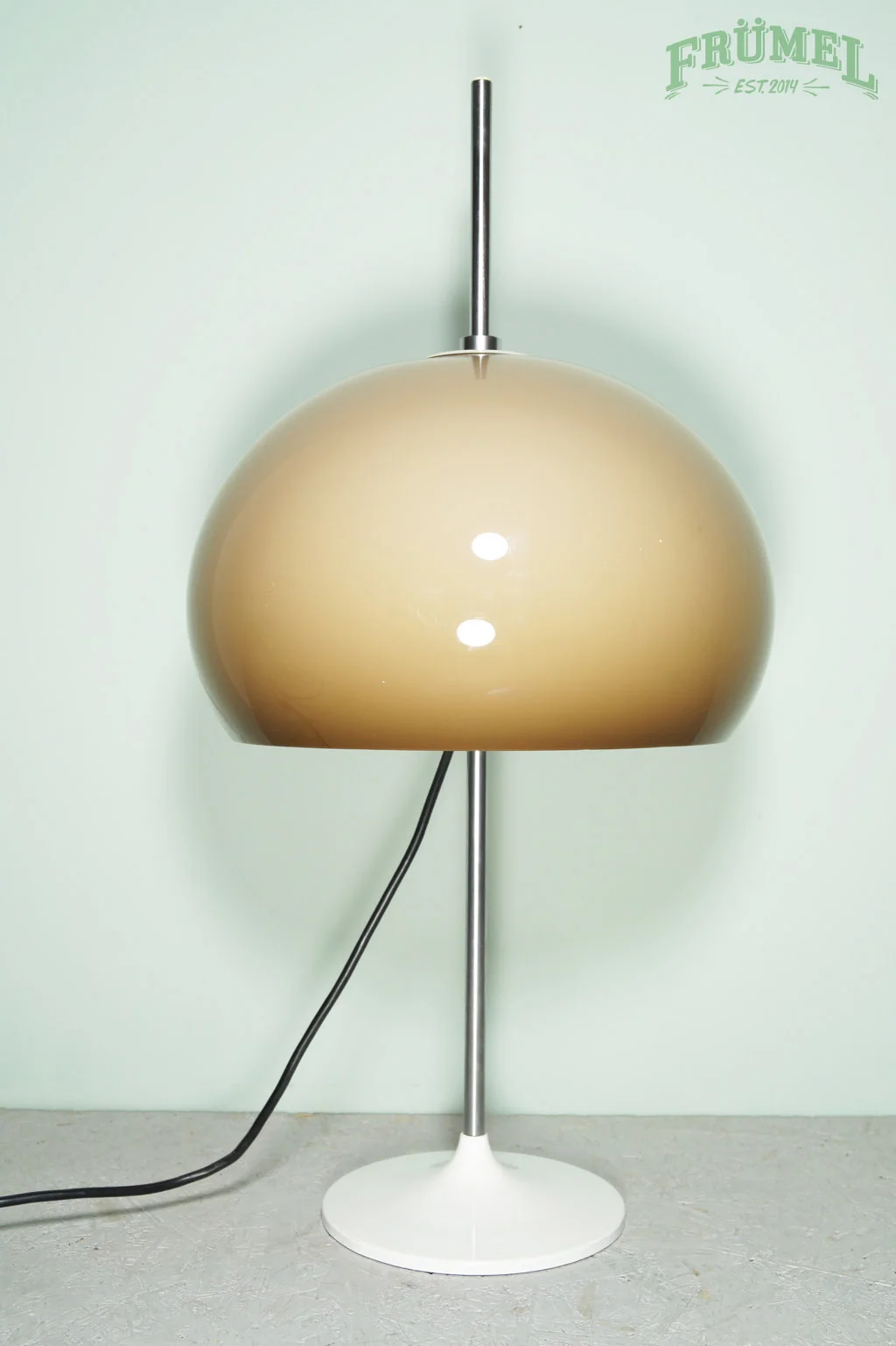 Een vintage vloerlamp met een grote, ronde kap, geplaatst op een lichte vloer tegen een pastelgroene muur, met een USB-type stekker. Het logo 'FRÜMEL' en de tekst 'EST. 2014' staan rechtsboven in de hoek.